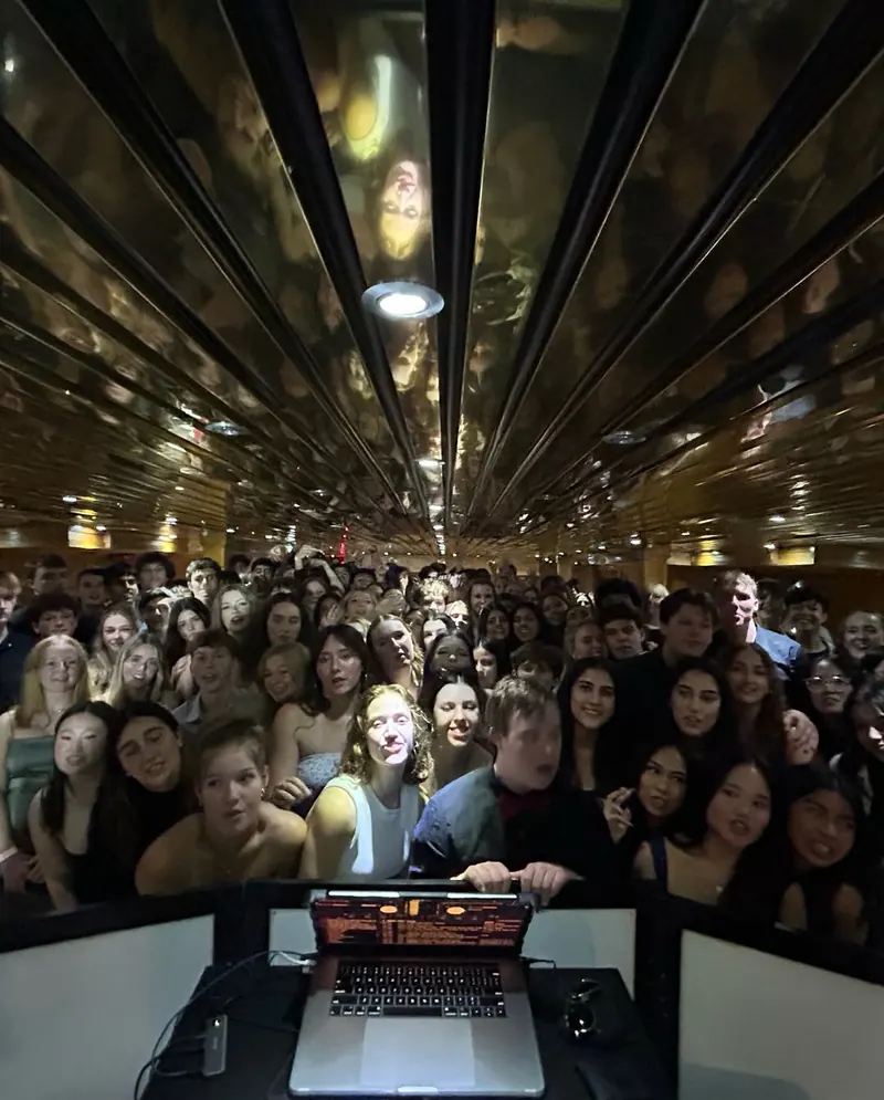 A crowd smiles toward the DJ laptop under a mirrored ceiling